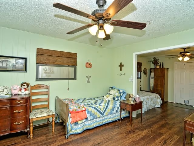A cozy senior living bedroom with a single bed covered in a blue and yellow quilt, a wooden chair, a small wooden dresser with stuffed animals on top, and a ceiling fan with lights. The room has light green walls and wooden flooring. There is an open doorway leading to another room with a bed and a ceiling fan visible.