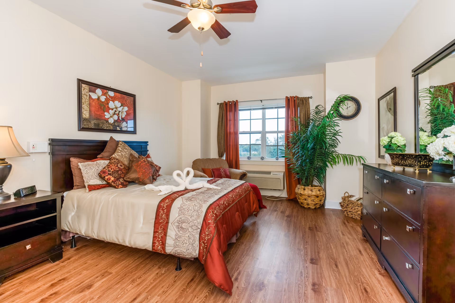 A neatly decorated bedroom with a made bed, dresser, window, potted plants, and ceiling fan.