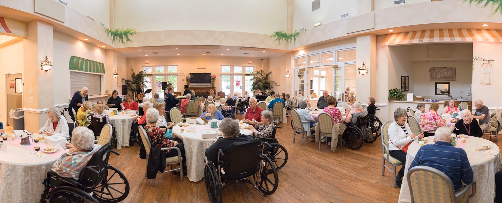 A large dining room filled with elderly residents seated at round tables covered with white tablecloths, enjoying meals and socializing. The room has high ceilings, large windows letting in natural light, and warm, inviting decor with plants and wall sconces. Some residents are in wheelchairs, and staff members are assisting them.