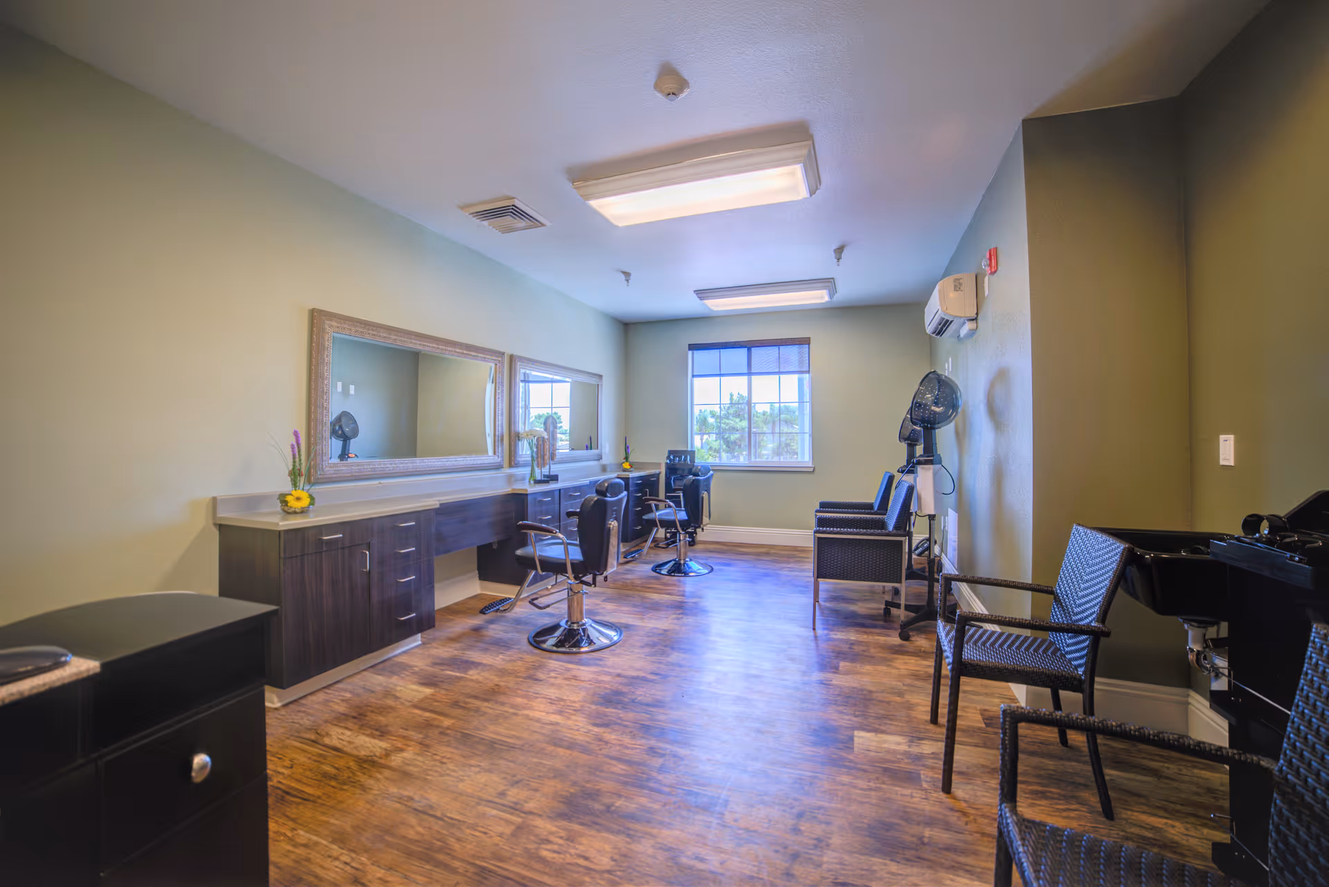 Interior view of a salon room with wooden flooring, two styling chairs in front of a long counter with large mirrors, a window letting in natural light, and additional seating along the right wall including a hair dryer station.