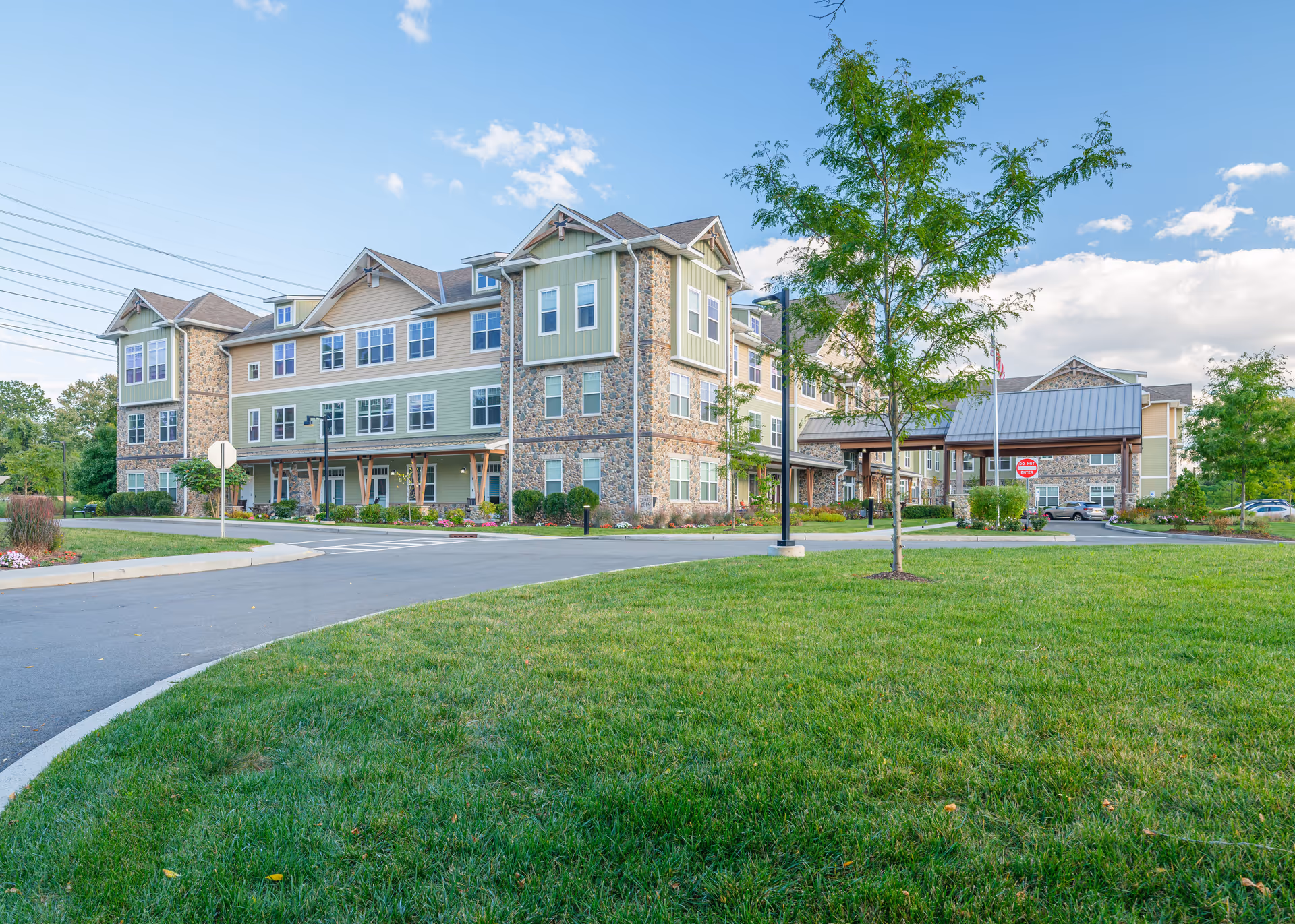 Exterior view of a multi-story senior living facility building with stone and siding facade, surrounded by green grass, trees, and a driveway under a partly cloudy sky.