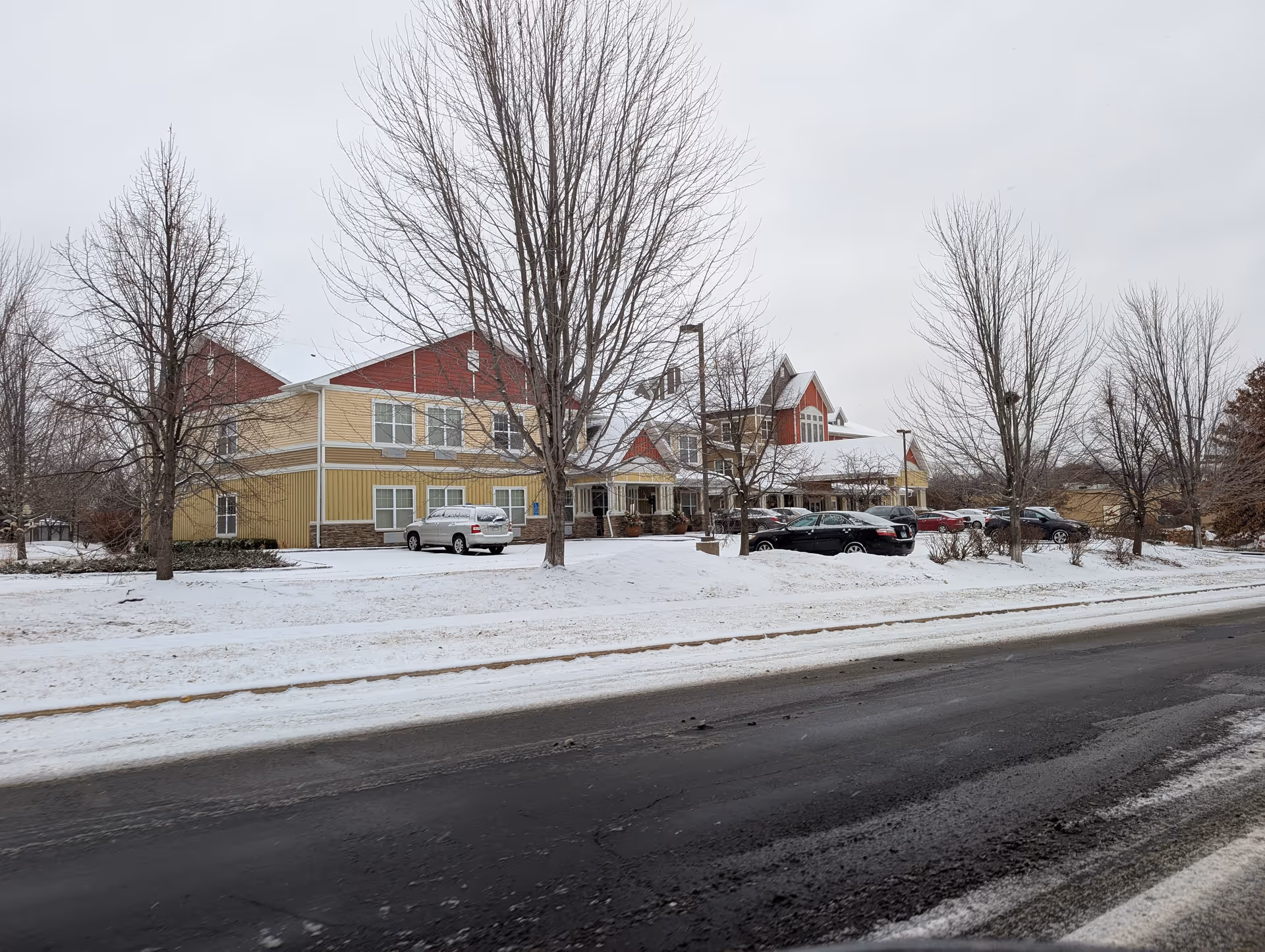 Exterior front view of a senior living building in winter with snow on the ground, parked cars, and leafless trees.