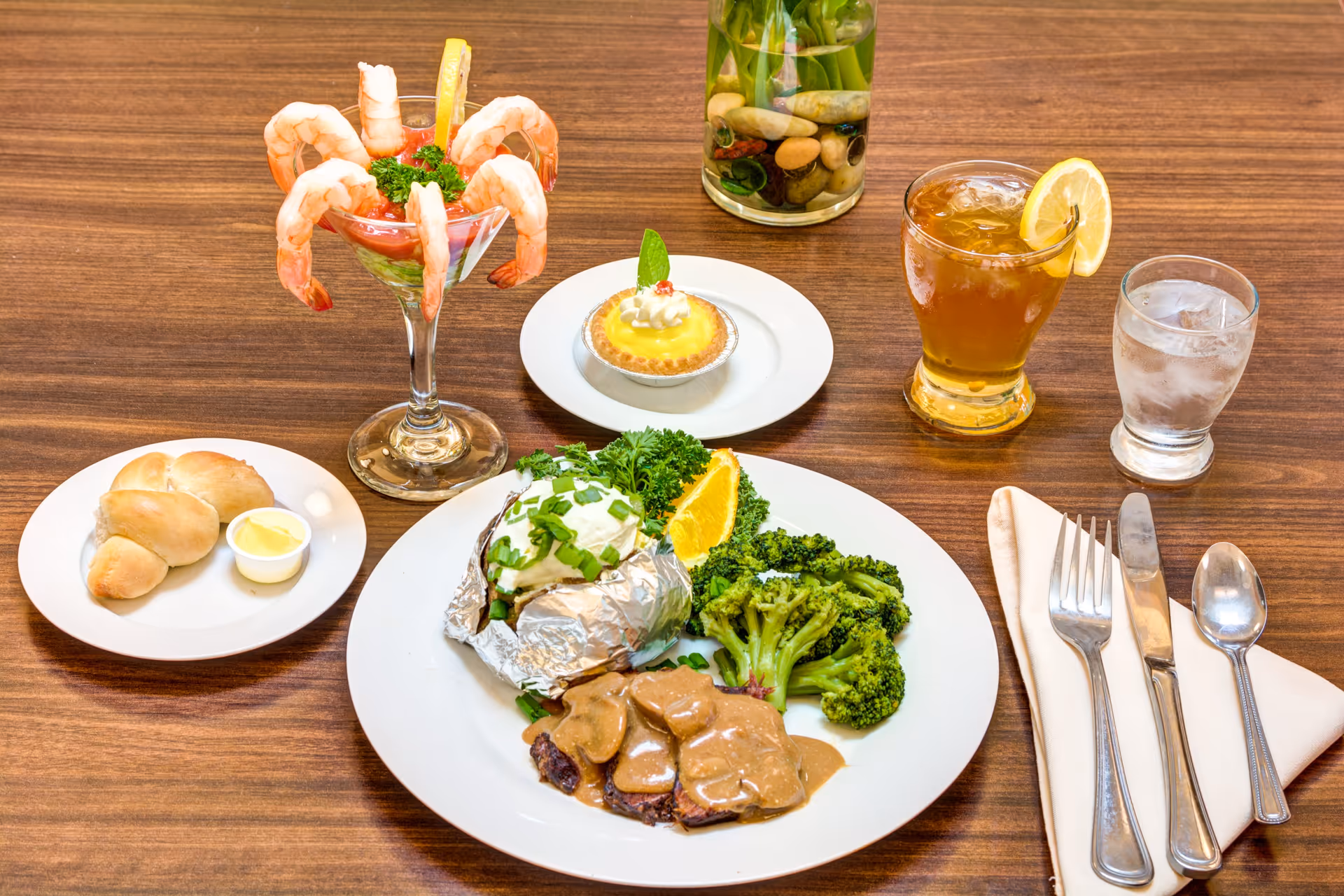 A meal served on a wooden table featuring a plate with sliced meat covered in gravy, steamed broccoli, a baked potato topped with sour cream and chives, and an orange slice. There is also a small plate with two bread rolls and butter, a shrimp cocktail in a glass with cocktail sauce and lemon garnish, a small lemon tart dessert on a white plate, a glass of iced tea with a lemon slice, and a glass of water. Silverware including a fork, knife, and spoon is placed on a folded napkin to the right.