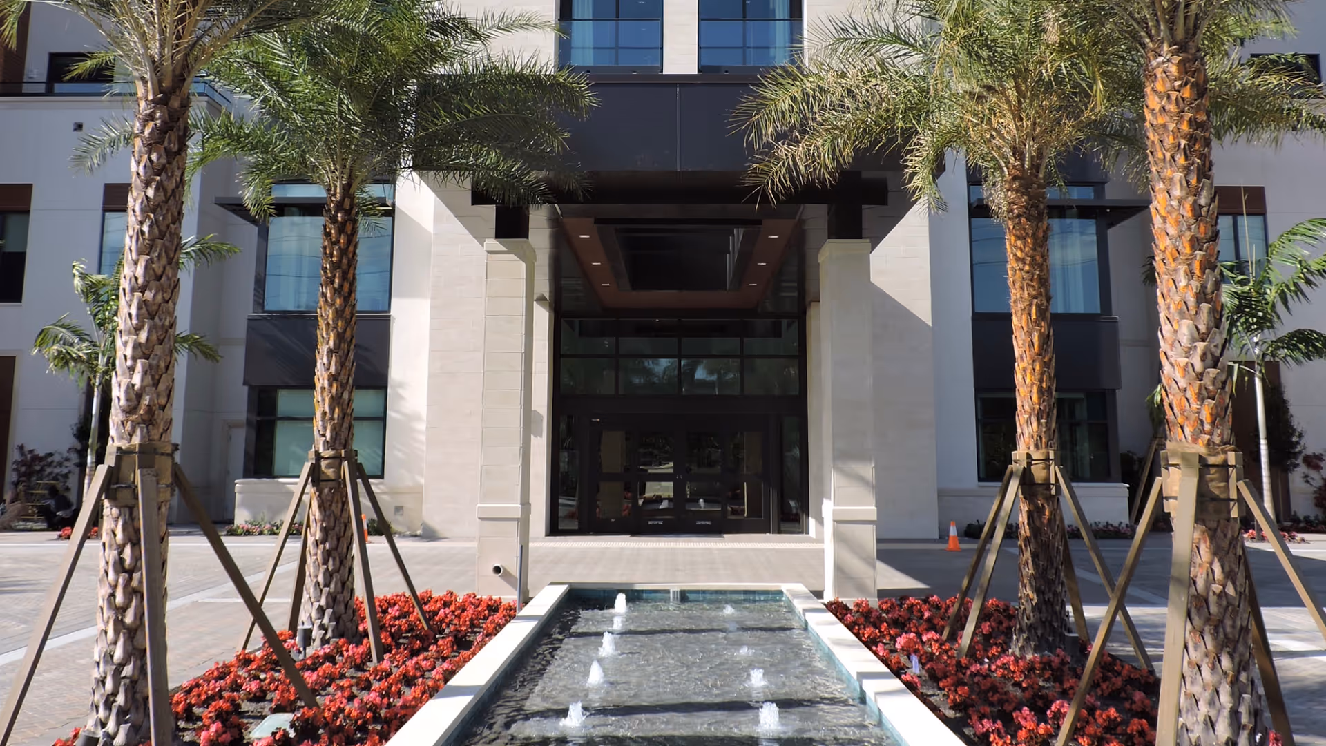 Entrance of a modern building with a water fountain in the foreground, flanked by palm trees and red flowers on both sides. The building has large windows and a covered entryway.