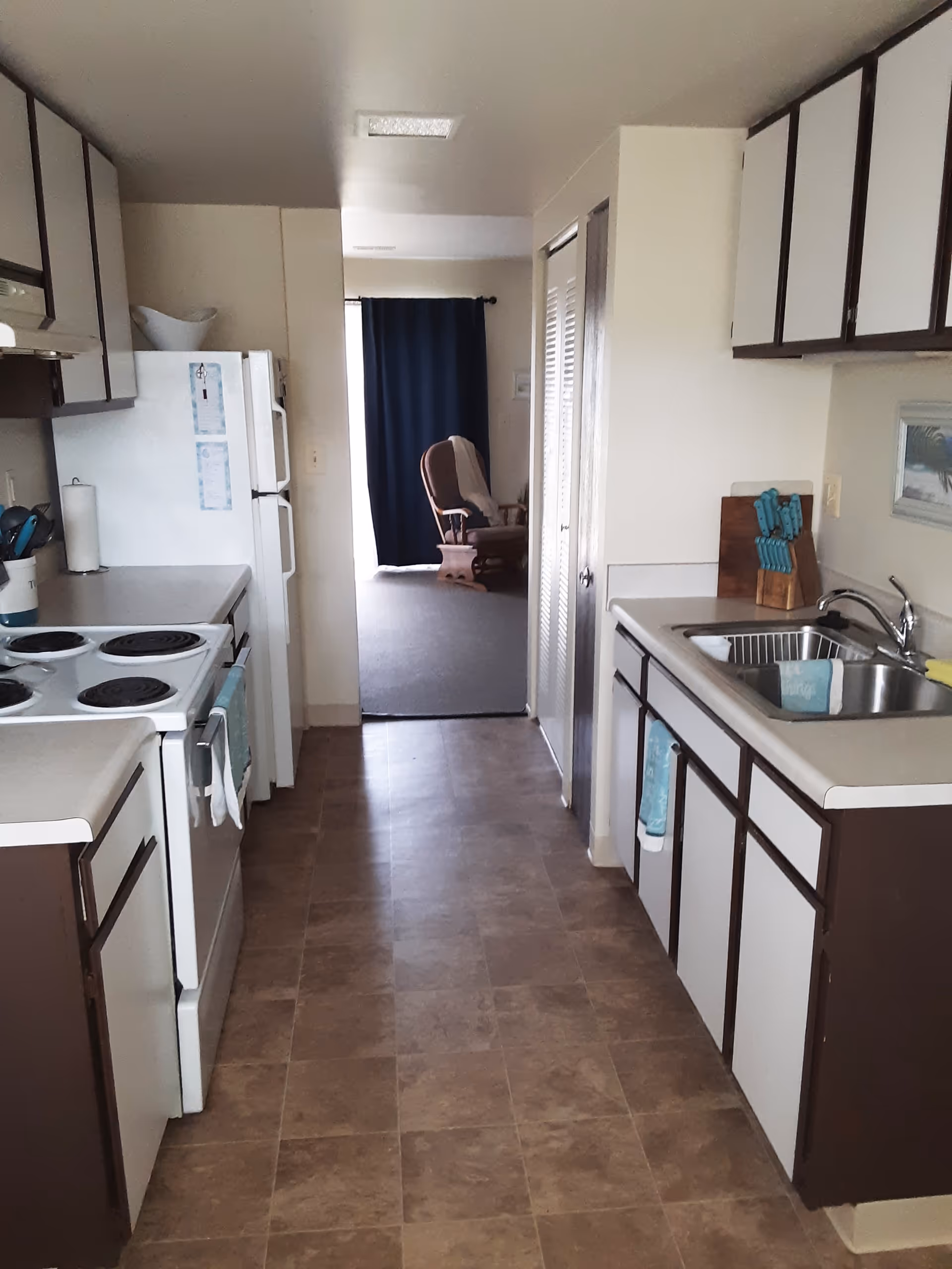 A narrow kitchen with brown and white cabinets on both sides, a white electric stove and refrigerator on the left, and a sink with a faucet on the right. There are kitchen utensils and a knife block on the counters. At the end of the kitchen, there is an open doorway leading to a room with a rocking chair and dark curtains.