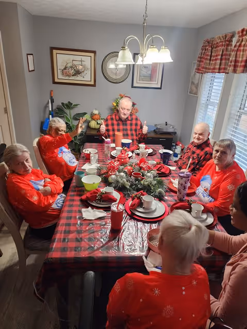 A group of elderly people sitting around a dining table decorated with a red and black checkered tablecloth and a festive centerpiece. Most of them are wearing red holiday-themed sweaters. The room has gray walls with framed pictures and a window with red plaid curtains. A chandelier with five lights hangs above the table.