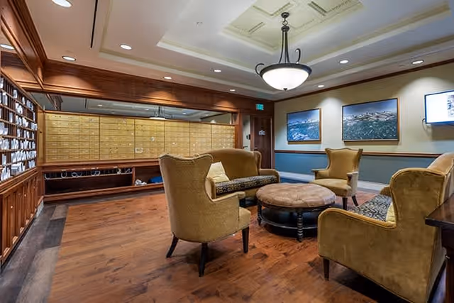 A cozy lounge area with mustard yellow upholstered chairs and a matching sofa arranged around a round ottoman. The room features wooden flooring, wood-paneled walls with built-in mailboxes, and framed aerial photographs on the walls. A ceiling light fixture hangs above, providing warm lighting.