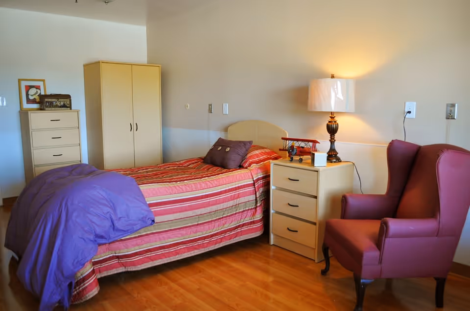 A senior living bedroom with a single bed covered in a red and beige striped bedspread and a purple blanket. Next to the bed is a wooden nightstand with a lamp, a small model airplane, and a white cube. There is a purple armchair to the right of the nightstand. In the background, there is a wooden wardrobe and a chest of drawers with a framed picture and a decorative box on top. The room has wooden flooring and light-colored walls.