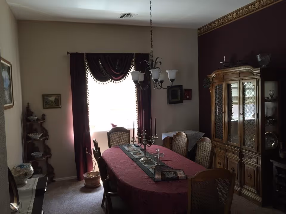 Formal dining room with a long table covered in a burgundy tablecloth, chandelier overhead, china cabinet, and a window with dark drapes.