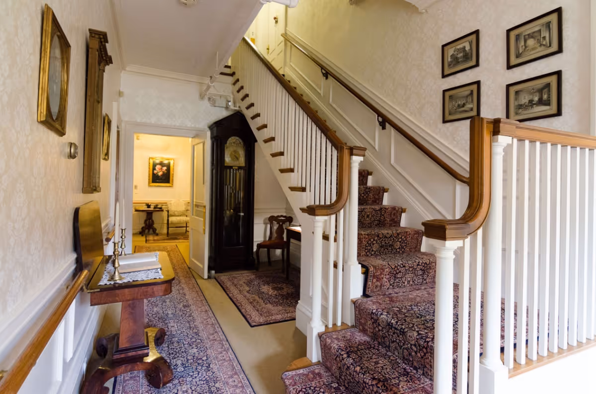 Interior view of a hallway in a senior living facility featuring a carpeted staircase with wooden handrails, an antique grandfather clock, framed artwork on the walls, and a small wooden table with decorative items. The hallway leads to another room with additional furniture and artwork.