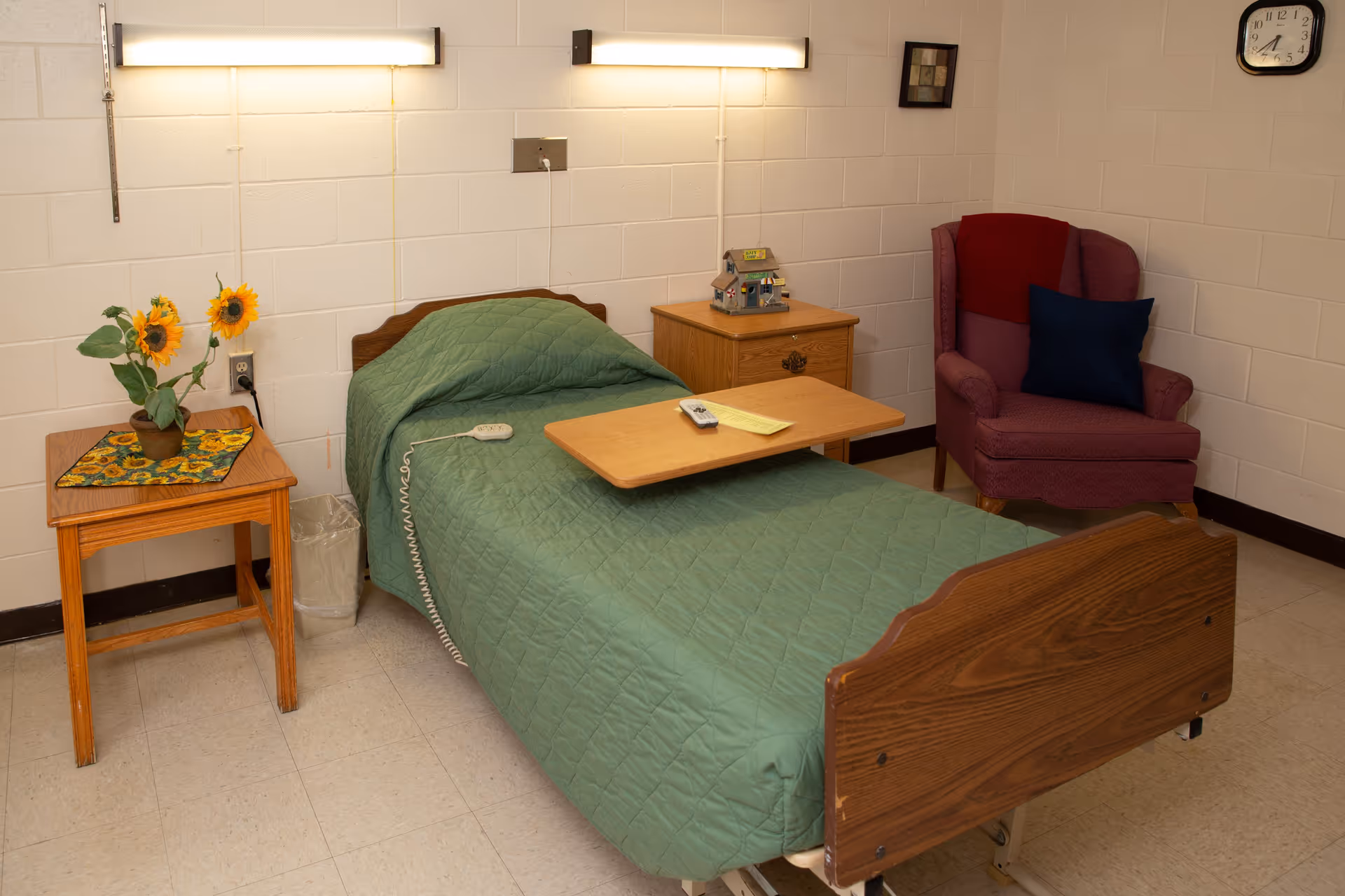 A small room with a single hospital-style bed covered with a green quilt. There is a wooden overbed table with a remote and a note on it. To the left of the bed is a wooden side table with a potted sunflower plant and a trash bin underneath. To the right of the bed is a wooden nightstand with a decorative house model on top. A red upholstered armchair with a dark blue pillow is in the corner. The walls are white cinder block with two fluorescent lights above the bed and a clock on the wall showing 3:10.