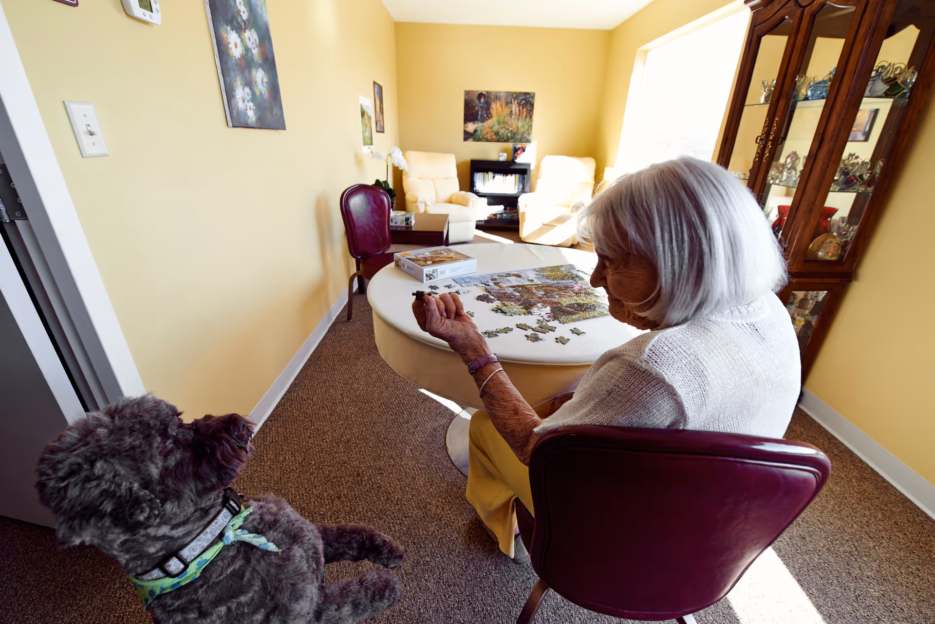 An elderly woman sitting at a round table working on a jigsaw puzzle in a cozy room with yellow walls. A small dog is sitting nearby looking up at her. The room has a glass display cabinet, two cream-colored armchairs, and framed pictures on the walls.