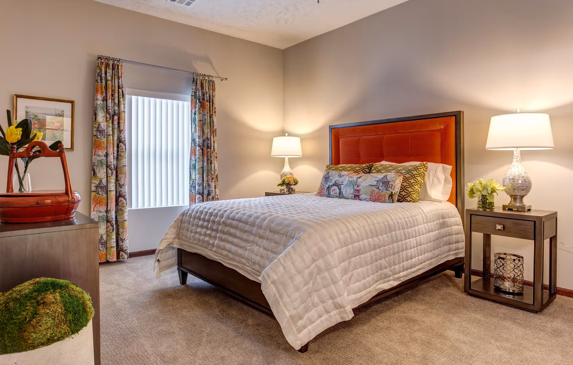 A cozy bedroom in Sky Pointe Retirement Resort featuring a bed with a red upholstered headboard, white quilted bedding, and decorative pillows. The room has beige walls, carpeted floor, two bedside tables with lamps, a window with vertical blinds and colorful curtains, a dresser with a red decorative tray and a framed picture on the wall.
