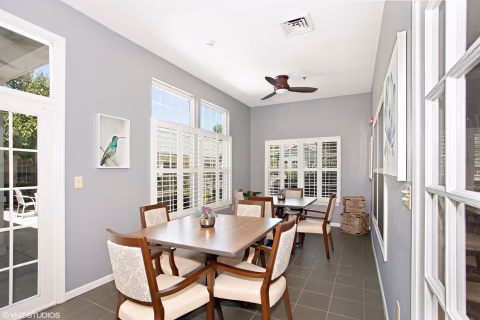 A bright dining room with two wooden tables and eight cushioned chairs. The room has gray walls, large windows with white shutters, and a ceiling fan. There are framed artworks on the walls and a small potted plant on each table. A glass door leads to an outdoor patio area with chairs.