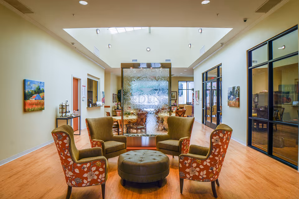 A bright and spacious common area in a senior living facility with four upholstered armchairs arranged around a round ottoman. The chairs have floral and solid patterns. Behind the seating area is a large decorative water feature with the word 'EDEN' visible on it. The room has wooden flooring, light-colored walls, several paintings, and glass-walled offices or rooms on the right side. Skylights and recessed lighting illuminate the space.