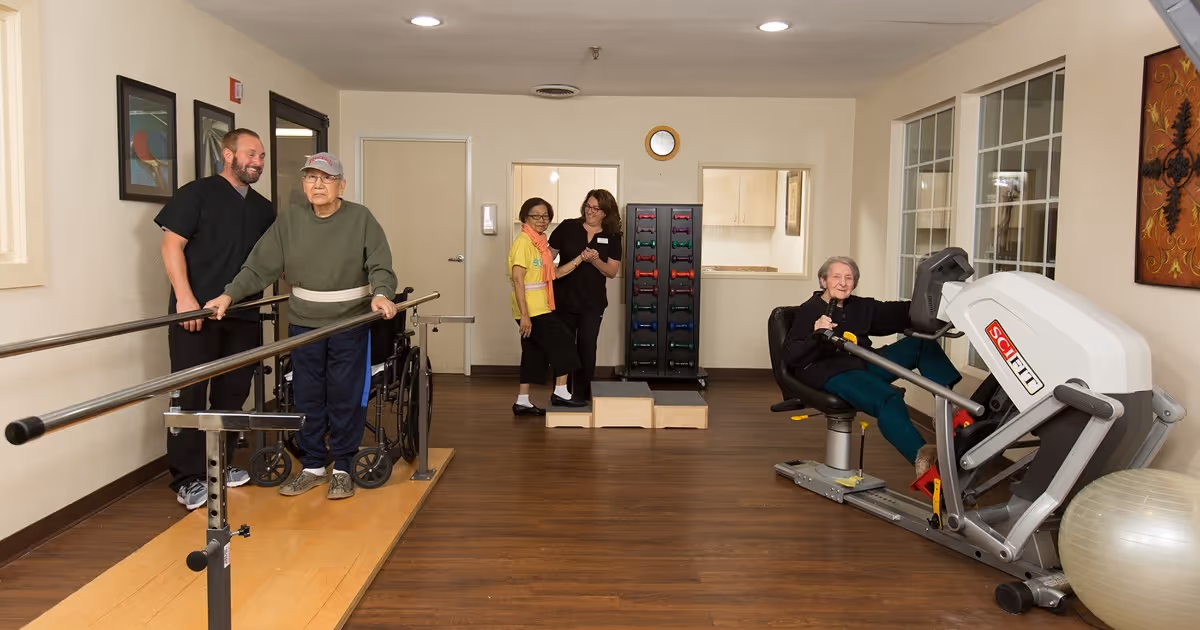An elderly man using parallel bars for walking assistance with a caregiver standing beside him, and an elderly woman exercising on a seated leg press machine in a rehabilitation or fitness room. Two other women and a caregiver are standing in the background near a rack of colorful dumbbells. The room has wooden flooring, light-colored walls, and framed artwork on the walls.
