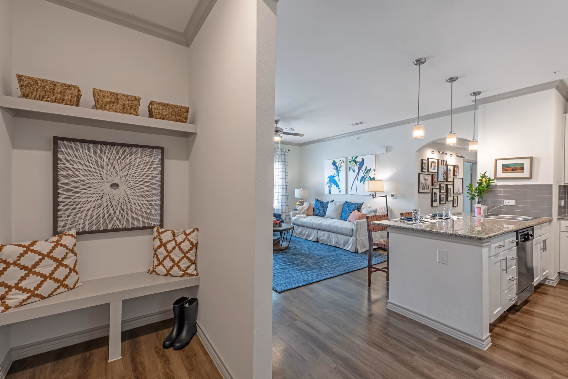 Open-plan interior showing an entryway bench with pillows and baskets leading into a living area with a sofa and blue rug and a kitchen island with pendant lights.