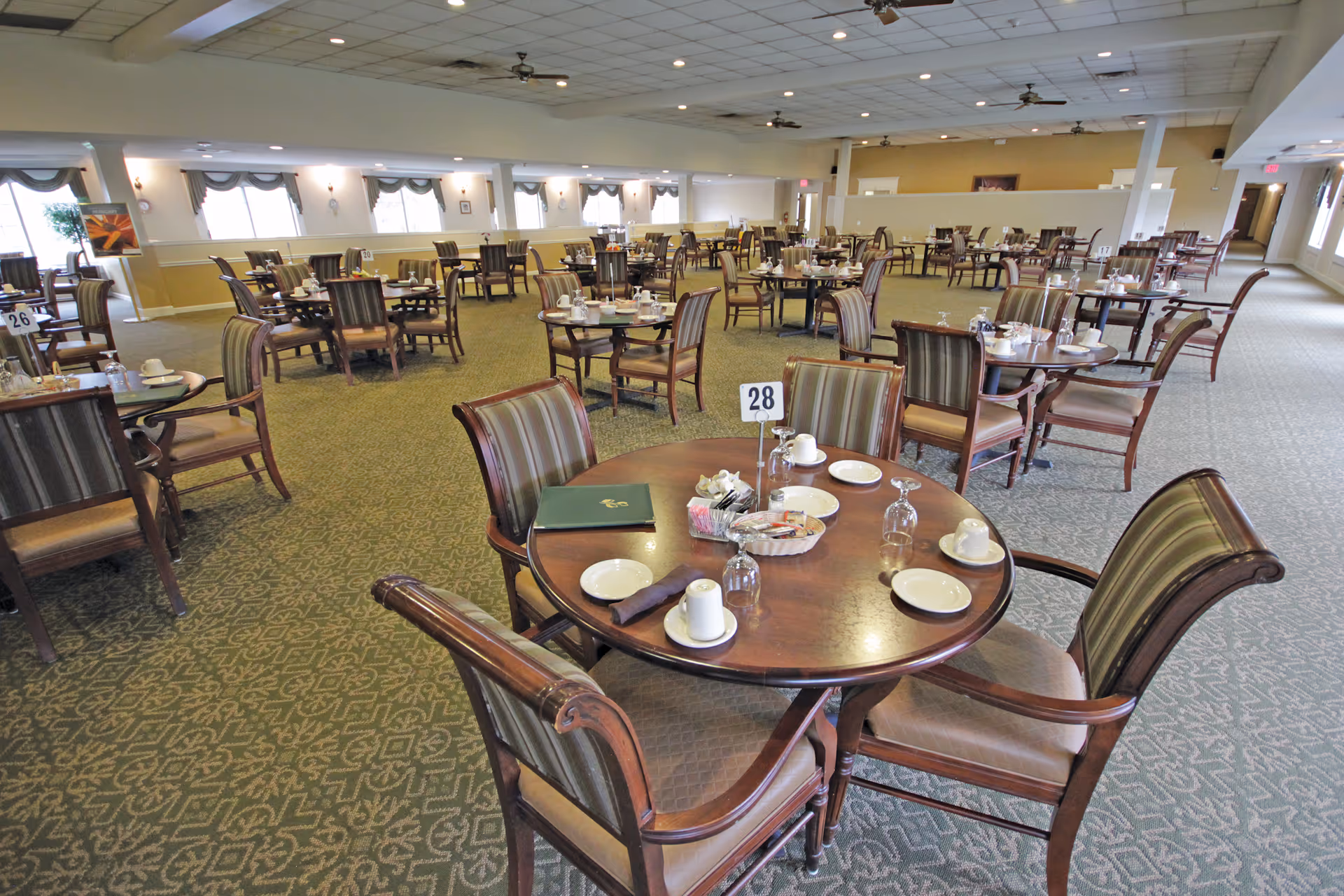 A spacious dining room with multiple round wooden tables and cushioned chairs arranged neatly. Each table is set with plates, cups, glasses, and napkins. The room has large windows with drapes, ceiling fans, and a patterned carpet floor.