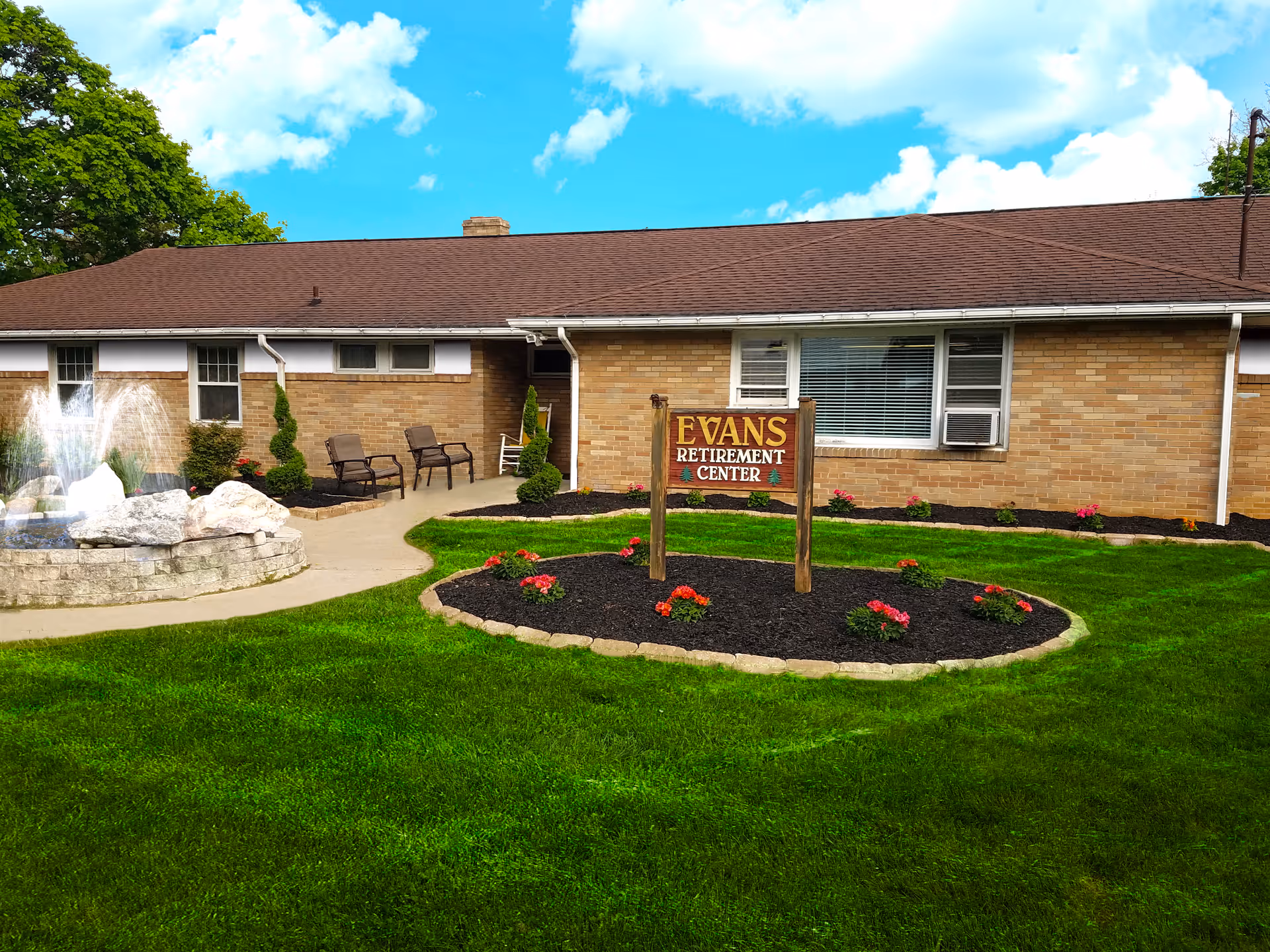 Exterior view of Evans Retirement Center showing a single-story brick building with a brown roof, a well-maintained lawn, flower beds with red flowers, a stone-bordered garden area, a water fountain on the left, and a wooden sign with the facility's name in front.