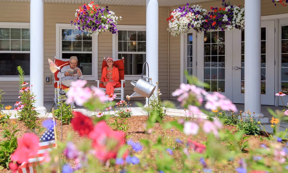 Two elderly women sitting on red cushioned rocking chairs on a porch with beige siding and white columns. There are hanging flower baskets above them and a garden with colorful flowers and an American flag in the foreground.