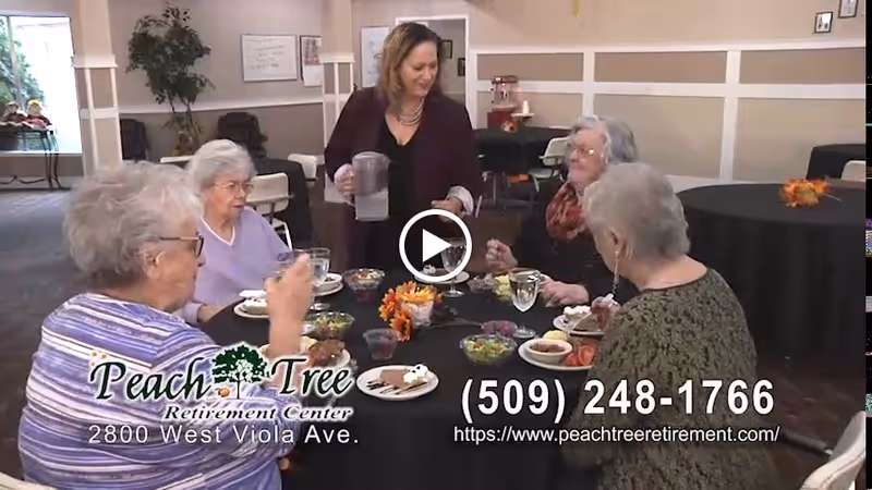 A group of elderly women sitting around a round table with black tablecloth, eating a meal together in a dining area. A staff member is standing and pouring water for one of the women. The room has beige walls and several other tables with chairs.