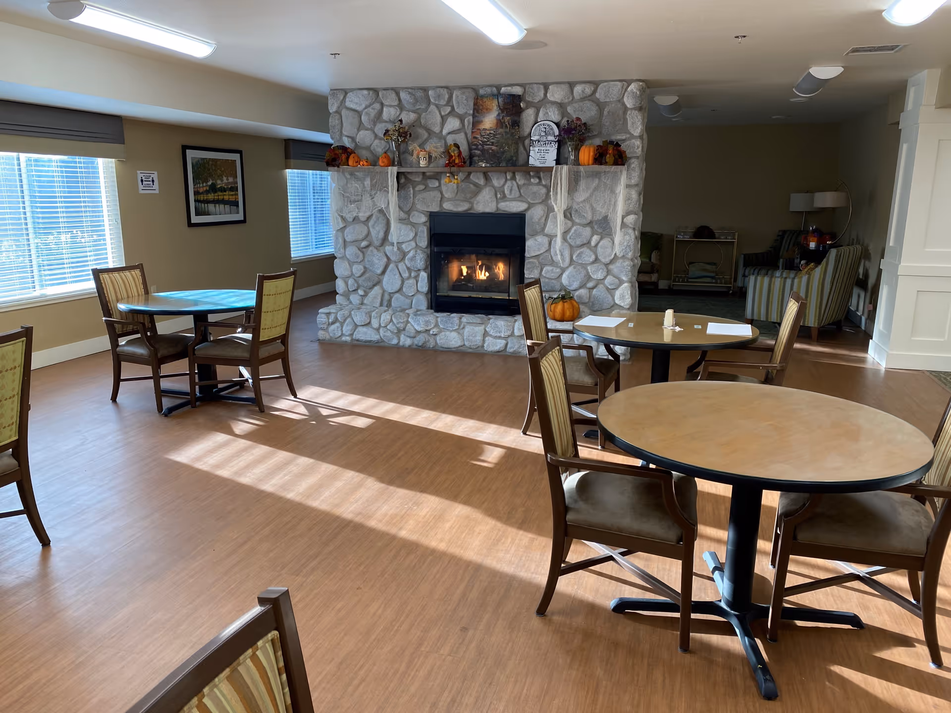 A bright common area with round wooden tables and chairs on a wood floor. A stone fireplace decorated with fall-themed items including pumpkins and faux cobwebs is centered in the room. Large windows with blinds allow natural light to fill the space. In the background, there are additional seating areas with armchairs and a lamp.