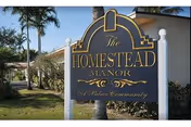 Outdoor view of a sign for The Homestead Manor, a palace community, with palm trees and a building in the background.