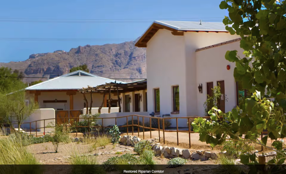 Single-story stucco building with a metal roof, desert landscaping and mountains in the background.