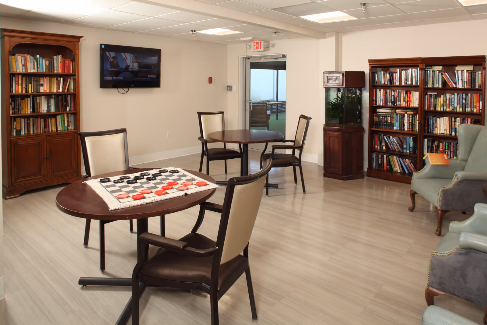 Bright common lounge with round tables and chairs, bookshelves, armchairs, a wall-mounted TV, and a checkers board on a table.