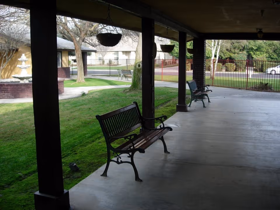 Covered outdoor patio with benches overlooking a grassy courtyard, fountain, and walkway.