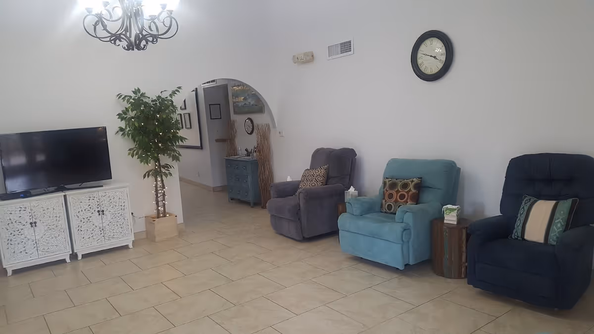 A cozy living room with three recliner chairs in gray, teal, and dark blue, each with decorative pillows. A small wooden side table with a tissue box is placed between two chairs. On the left side, there is a white ornate cabinet with a flat-screen TV on top and a potted plant with string lights. The room has beige tiled flooring, white walls, a black-framed clock, and a decorative chandelier hanging from the ceiling. An arched doorway leads to another area with a blue cabinet and wall decorations.