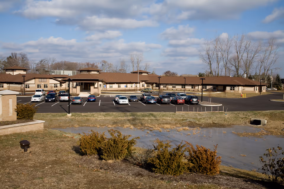 Exterior view of a single-story senior living facility building with a brown roof and beige walls. Several cars are parked in the parking lot in front of the building. The foreground includes a grassy area with some bushes and a small water retention pond. Leafless trees are visible in the background under a partly cloudy sky.
