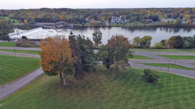 A scenic aerial view of a river with a small dam, surrounded by trees with autumn foliage and a grassy area in the foreground. Across the river, there are buildings and more trees extending into the distance under a cloudy sky.