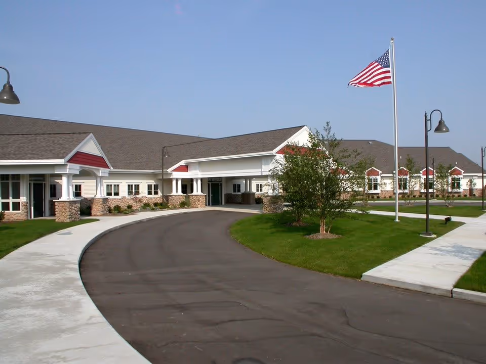 Exterior view of a single-story building with a curved driveway, green lawn, trees, and an American flag on a flagpole under a clear blue sky.
