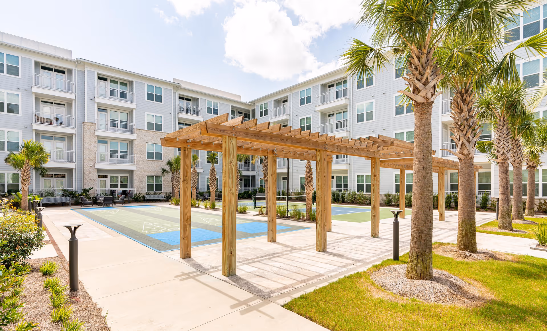 Outdoor courtyard area of a senior living facility with a wooden pergola, palm trees, and a shuffleboard court. The building surrounding the courtyard has multiple floors with balconies and large windows. The sky is partly cloudy.