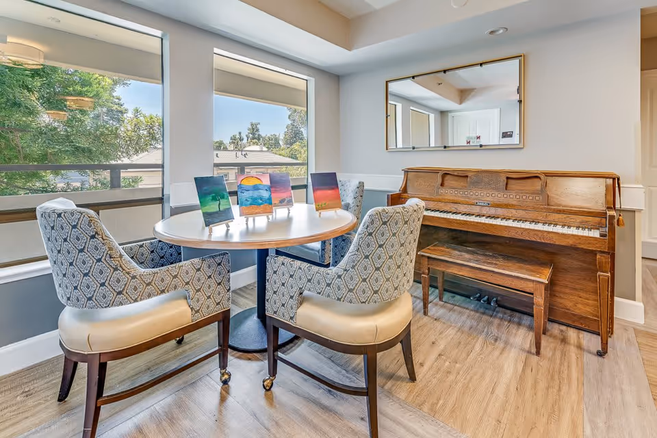 A cozy sitting area in a senior living facility with a round wooden table surrounded by four upholstered chairs. On the table are four small paintings displayed on stands. Behind the table is an upright wooden piano with a matching bench. A large rectangular mirror hangs on the wall above the piano. Two large windows provide natural light and a view of trees and rooftops outside.