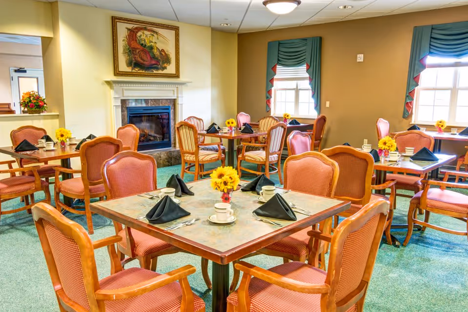 A cozy dining room with several wooden tables and cushioned chairs arranged neatly. Each table is set with black folded napkins, white cups and saucers, and small vases holding yellow flowers. The room features a fireplace with a framed painting above it, green carpet, and windows with green and red curtains allowing natural light to enter.