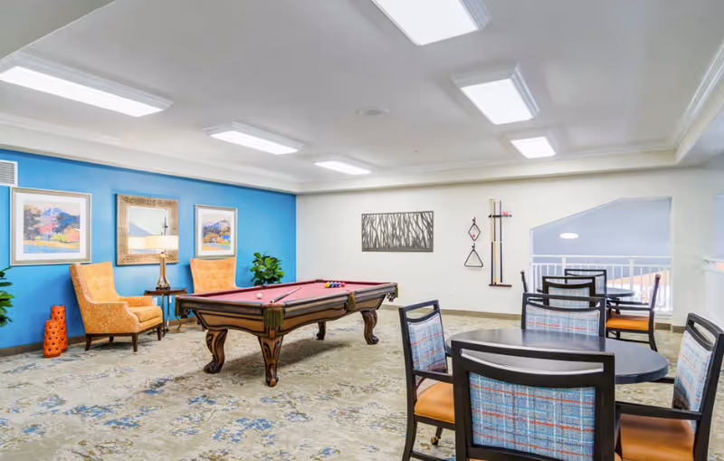 A bright and spacious game room with a pool table in the center, two orange armchairs with a side table and lamp against a blue accent wall, framed artwork, a decorative wall piece, and several tables with chairs arranged for seating.
