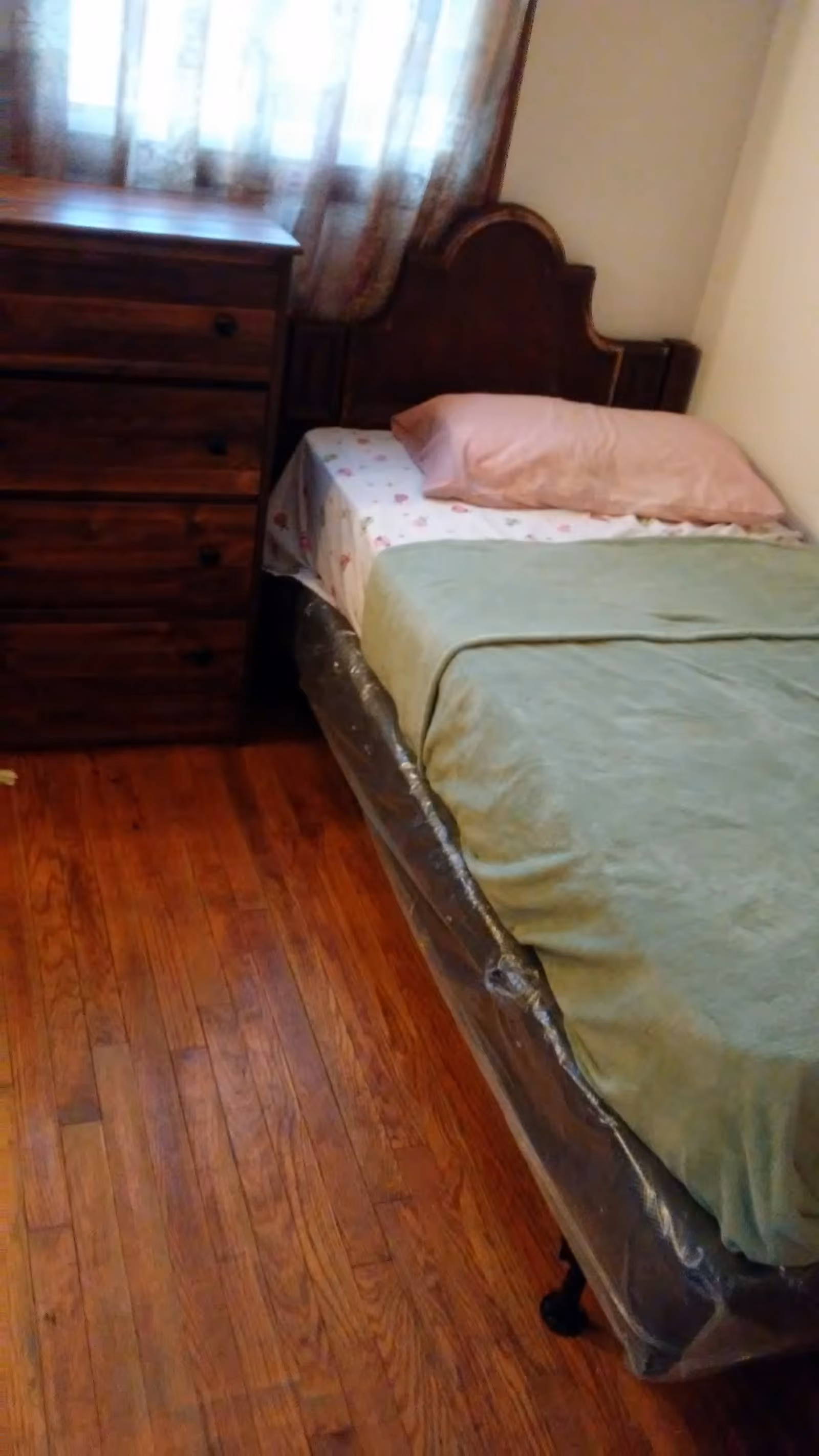 A small bedroom with a single bed covered with a green blanket and a pink pillow. Next to the bed is a wooden dresser with four drawers. A window with sheer curtains is behind the dresser, and the floor is made of polished wood.