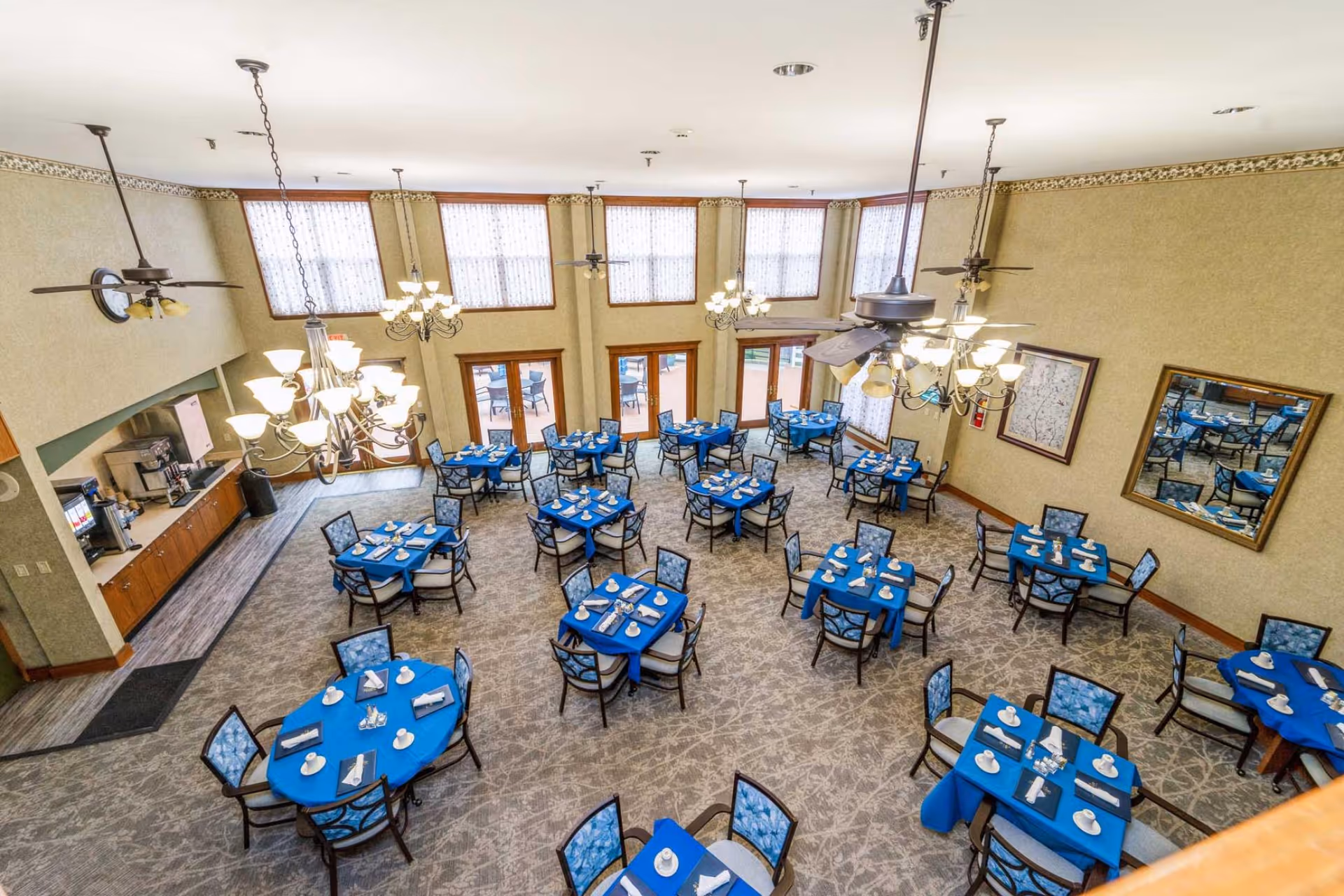 A spacious dining room with multiple round tables covered in blue tablecloths, each set with white cups, napkins, and silverware. The room has large windows with sheer curtains, ceiling fans, and chandeliers providing light. There is a coffee and beverage station along one wall, and framed artwork and a mirror on the opposite wall.
