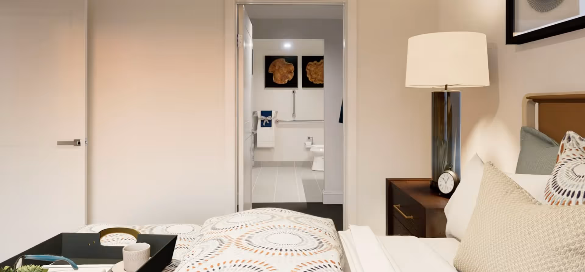 View of a bedroom with a bed featuring patterned bedding and pillows, a bedside table with a lamp and a clock, and an open door leading to a bathroom with a toilet, towel rack, and wall art.