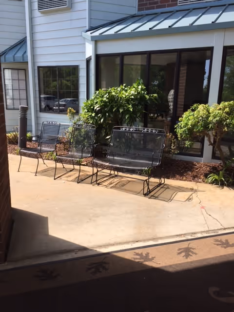 Outdoor seating area with three metal chairs and a metal bench on a concrete patio next to a building with large windows and some green bushes.