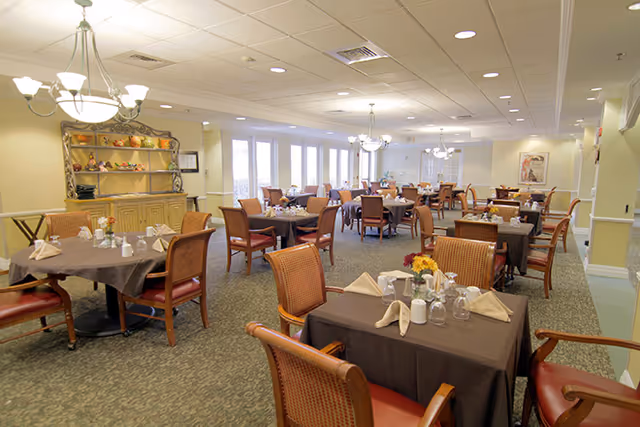 A spacious dining room with multiple tables covered in dark tablecloths, each set with folded napkins, glasses, and small flower arrangements. The room features comfortable wooden chairs with red cushions, carpeted flooring, and soft lighting from chandeliers. A decorative cabinet with shelves holding various items is visible against one wall, and large windows allow natural light to enter the space.