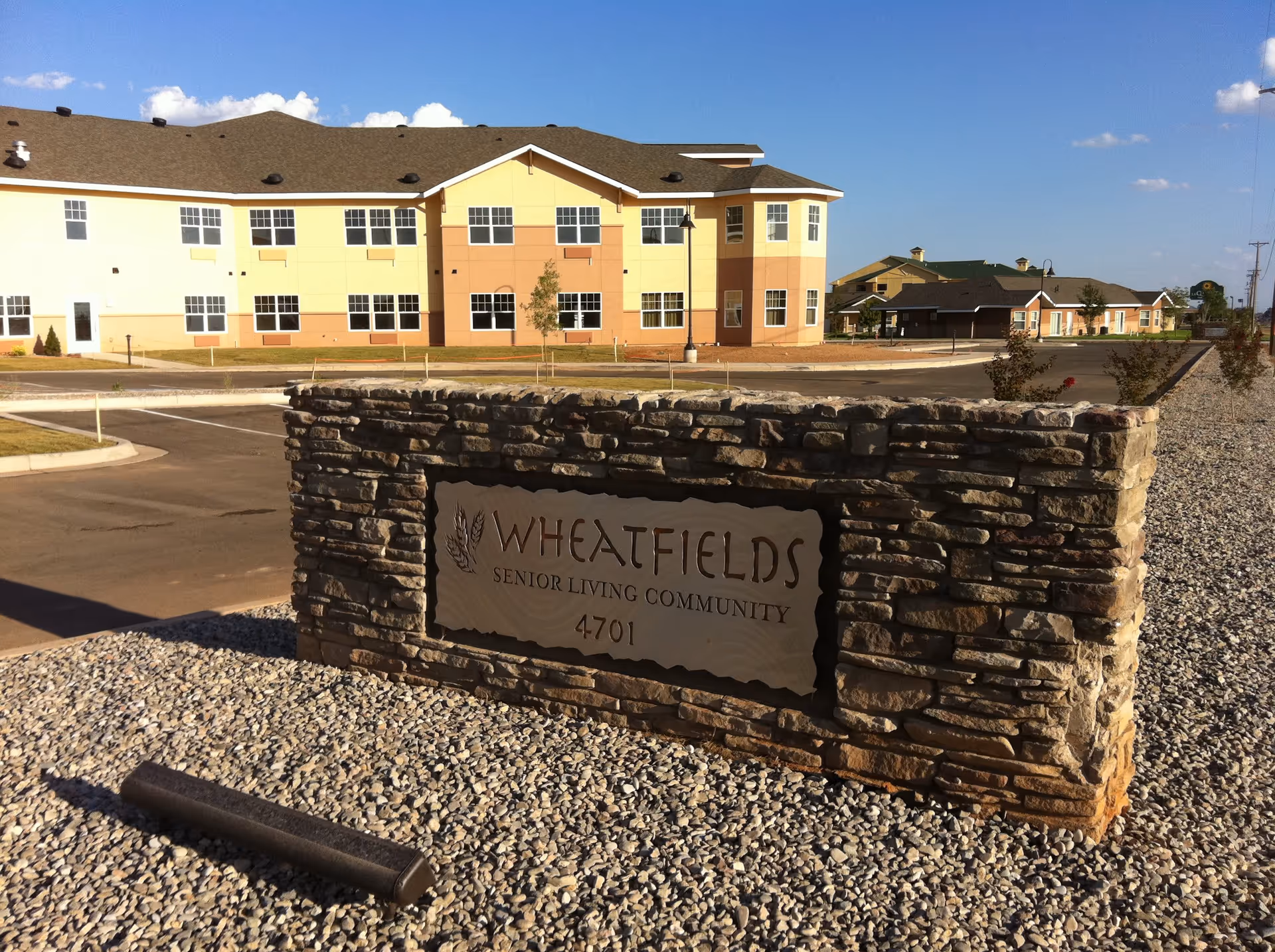 Stone sign with the text 'Wheatfields Senior Living Community 4701' in front of a large two-story senior living building under a blue sky with some clouds.