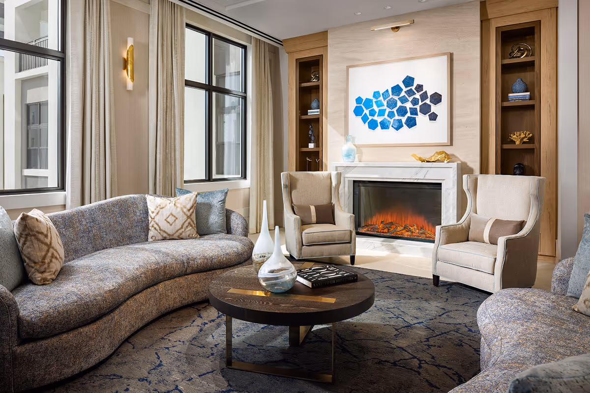 A cozy senior living room with a curved blue-gray sofa, two beige armchairs, a round wooden coffee table with decorative items, a fireplace with a modern electric fire, built-in wooden shelves with decorative objects, large windows with beige curtains, and a blue abstract artwork above the fireplace.