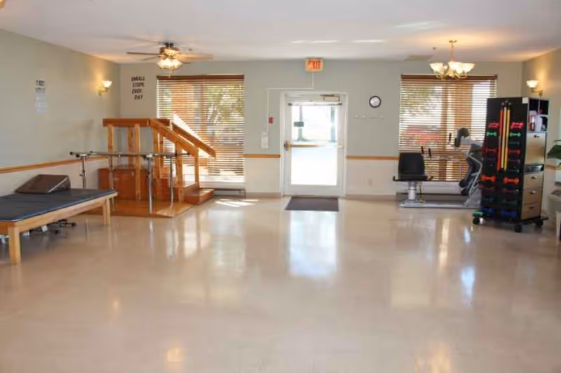 A spacious rehabilitation room with exercise equipment including a parallel bar setup, a therapy bed, a stationary bike, and a rack of colorful dumbbells. The room has large windows with blinds, a ceiling fan, and a door leading outside.