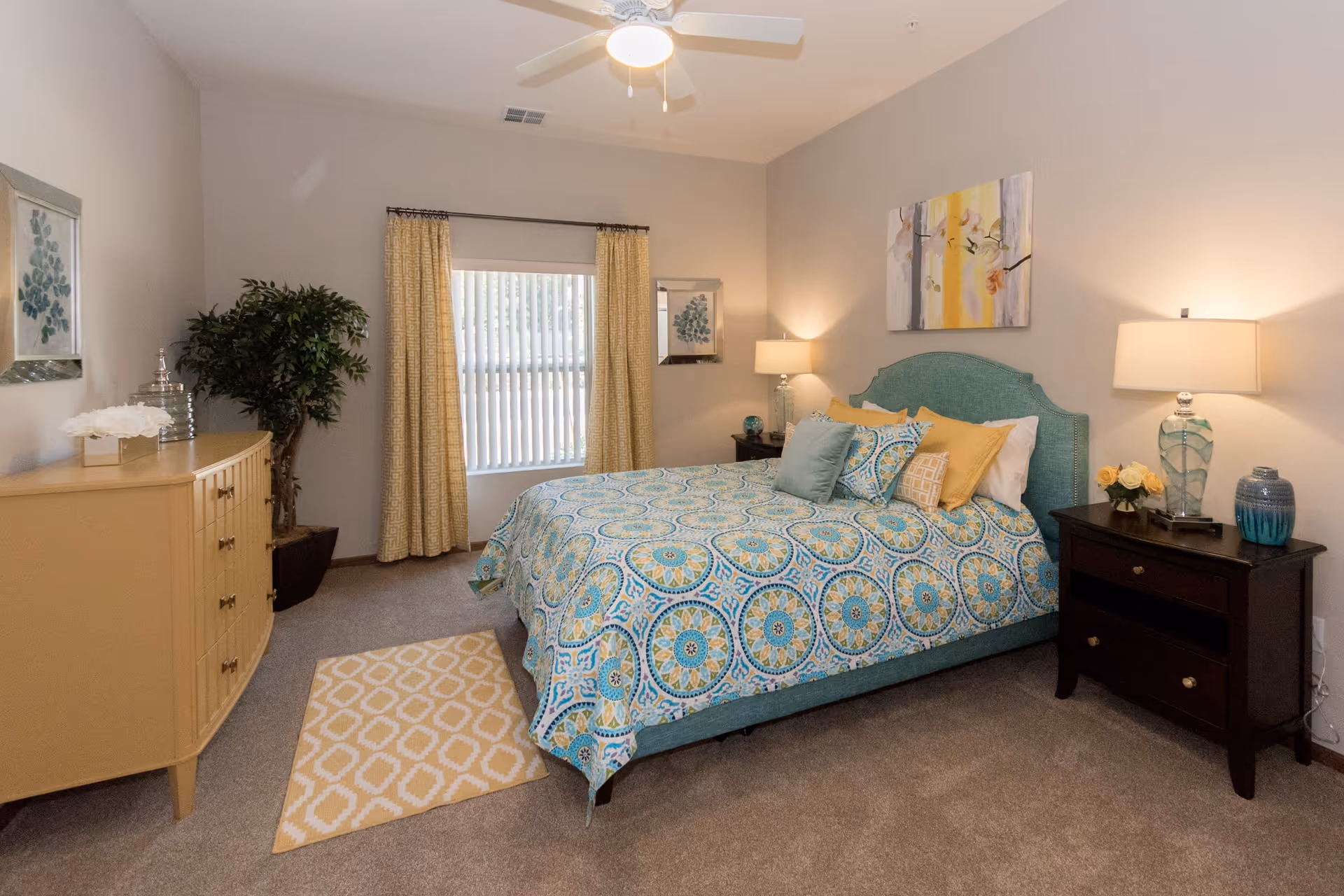A cozy bedroom with a patterned blue and yellow bedspread on a bed with a teal upholstered headboard. The room features beige walls, a ceiling fan with light, a window with yellow curtains, a beige dresser with decorative items, a potted plant, two dark wood nightstands with lamps and decorative vases, and a small yellow patterned rug on the carpeted floor.