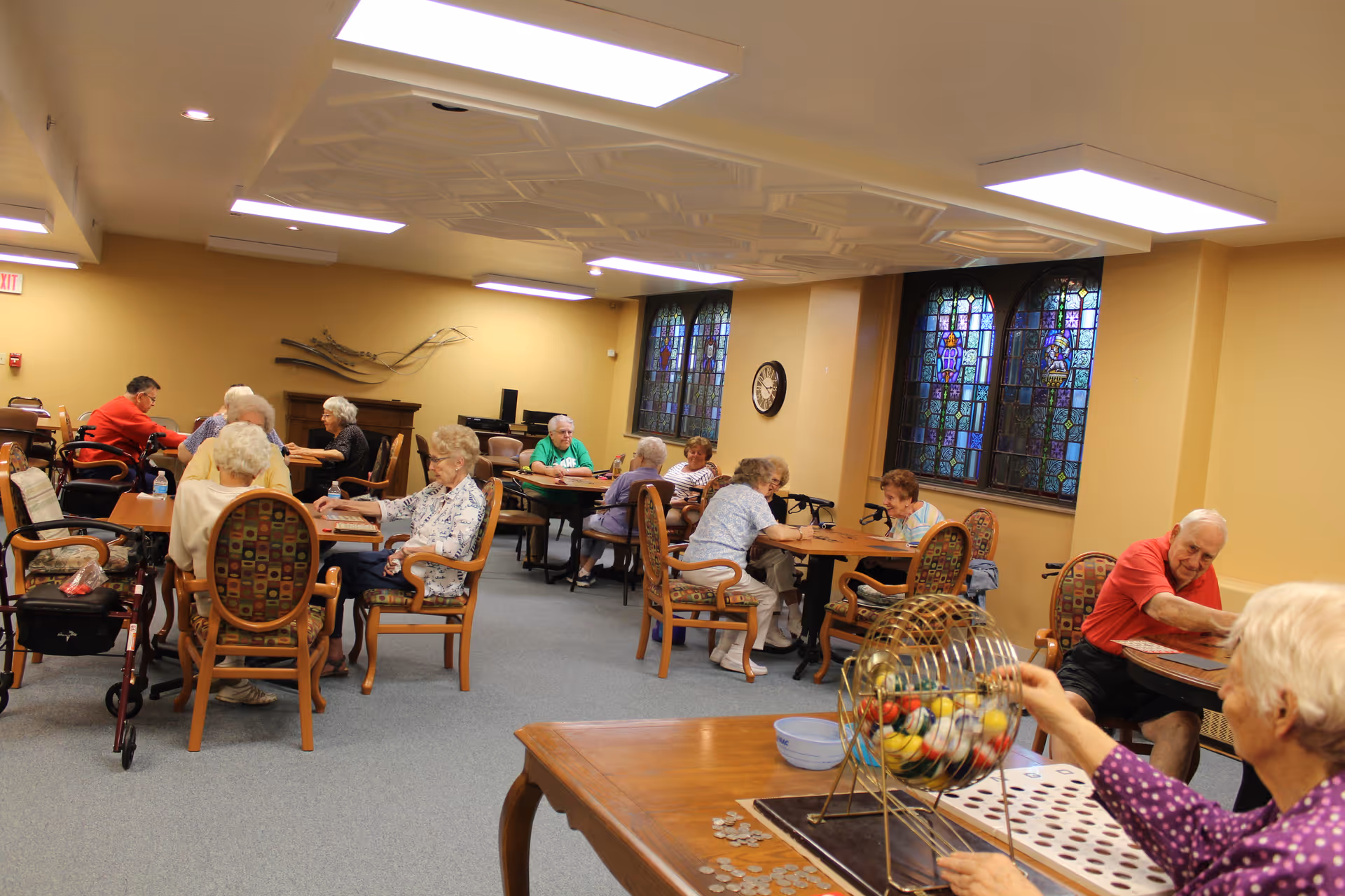 A group of elderly people sitting at tables in a well-lit room with stained glass windows, playing bingo. One person in the foreground is turning a bingo cage with numbered balls inside. The room has beige walls, patterned chairs, and a clock on the wall.
