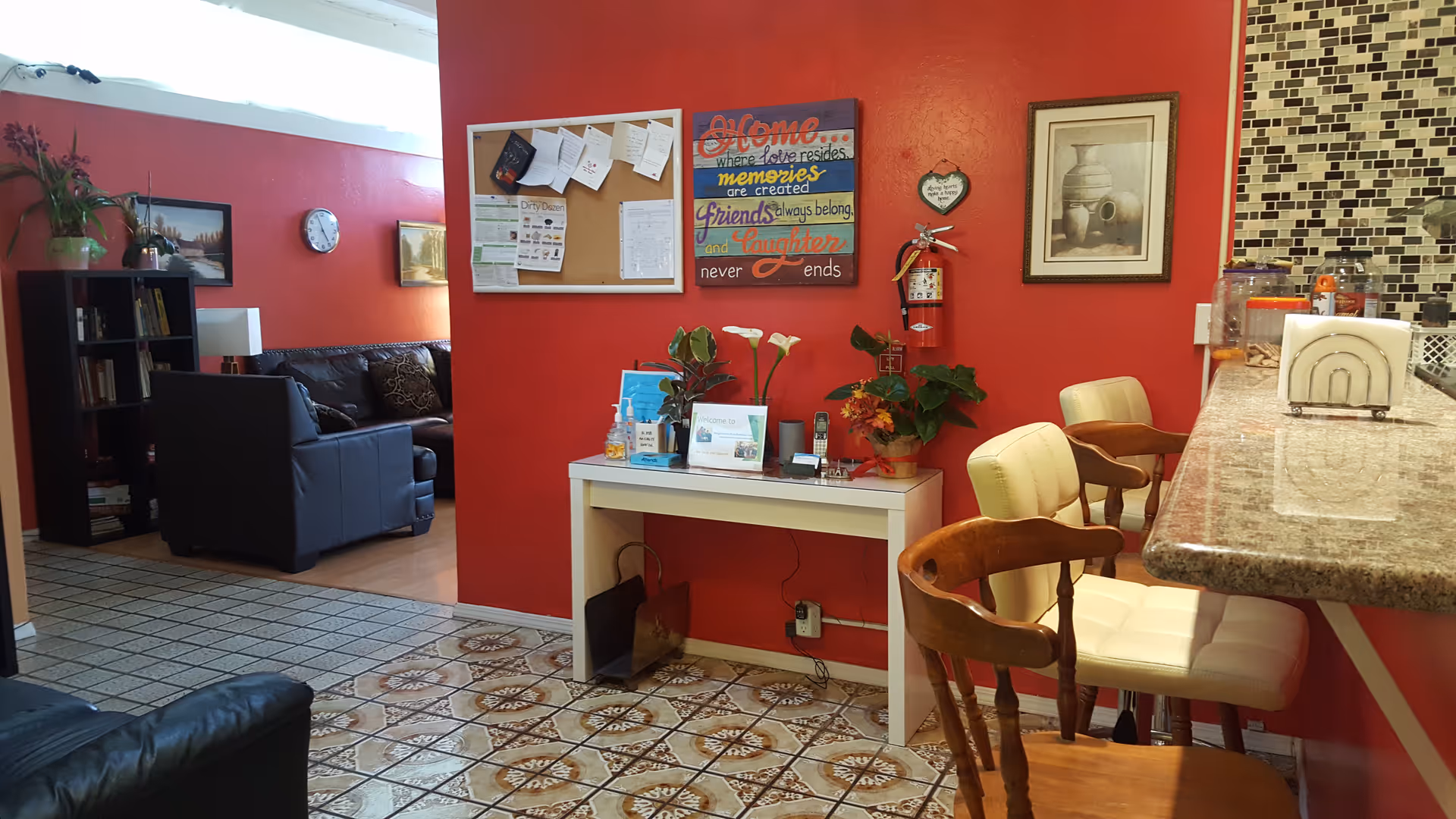 Interior view of a residential care facility showing a cozy common area with red walls. There is a white table with plants and informational materials against the wall, above which hangs a colorful sign with an inspirational quote. To the right, there is a granite countertop with two cushioned bar stools and a wooden chair. In the background, a living room area with black leather sofas, a bookshelf, and framed pictures on the walls is visible.