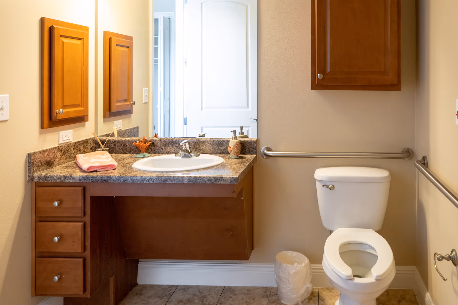 A clean bathroom with a white toilet featuring a raised seat and grab bars on both sides. There is a wooden vanity with a granite countertop, a white sink, a soap dispenser, and a pink towel. A mirror and wooden cabinet are mounted on the wall above the sink. The floor is tiled, and a small trash can is placed next to the toilet.